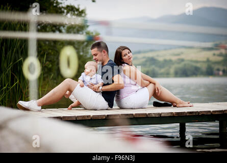 Il padre e la madre con bambino seduto sul molo in riva al lago Foto Stock