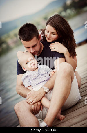 Il padre e la madre con bambino seduto sul molo in riva al lago Foto Stock