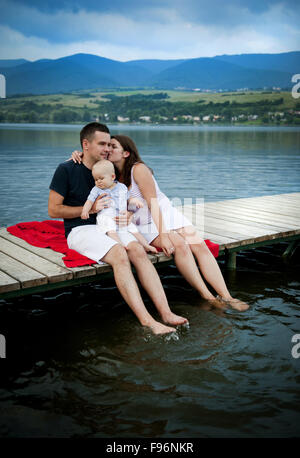 Il padre e la madre con bambino seduto sul molo in riva al lago Foto Stock