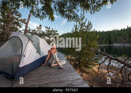 Un camper si rilassa su una piattaforma accanto alla sua tenda al tramonto. Curme West Island, la desolazione del suono del parco marino, British Columbia, Foto Stock