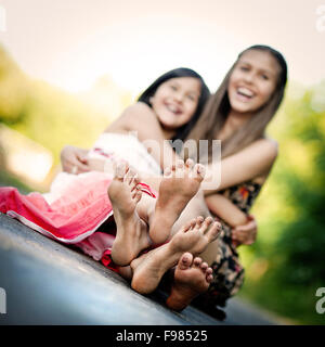 Due sorelle di ridere e abbracciando sul marciapiede nel verde parco di sole Foto Stock