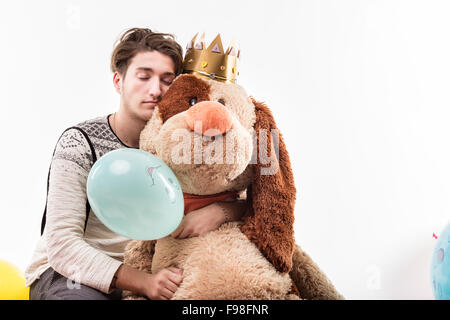 Giovane uomo abbraccia una grande bambola cane dorme da una parte forse avere una sbornia, il cane ha una corona e ci sono palloncini intorno Foto Stock