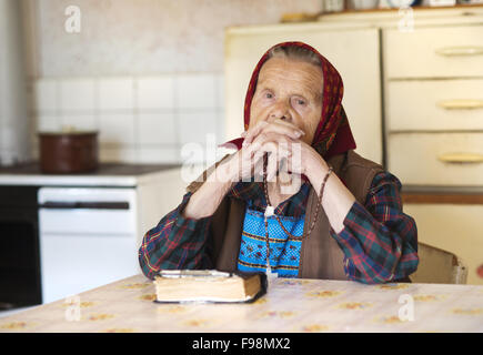 Molto vecchia donna indossa foulard di testa sta pregando nel suo paese La cucina di stile Foto Stock