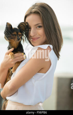 Giovane e bella donna che indossa abiti eleganti holding piccolo cane mentre passeggiate in estate park Foto Stock
