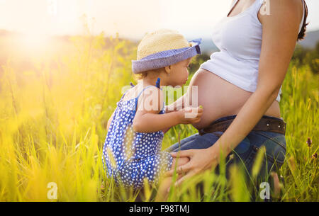 Carino bambina abbracciando e baciando la madre incinta pancia in estate la natura Foto Stock