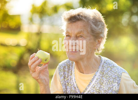 Ritratto di donna senior in grembiule apple mangiare nel giardino soleggiato Foto Stock