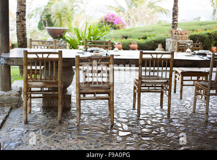 Tavolo con sedie sul patio in un giardino tropicale con alberi esotici e boccole Foto Stock