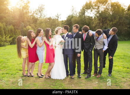 A piena lunghezza Ritratto di sposa giovane kissing in posa con damigelle e groomsmen nel verde parco di sole Foto Stock