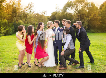 A piena lunghezza Ritratto di sposa giovane kissing in posa con damigelle e groomsmen nel verde parco di sole Foto Stock