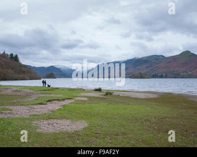 Due persone e un cane a camminare sulla spiaggia di Derwentwater Lake District Cumbria Regno Unito su nuvoloso giorno. Foto Stock