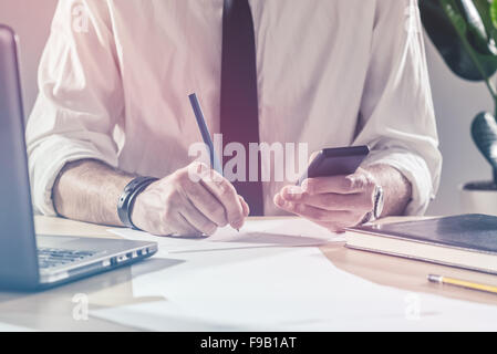 Imprenditore iscritto note dal telefono cellulare, lavoro straordinario alla scrivania in ufficio, il multitasking e la scadenza di un progetto concetto, Foto Stock