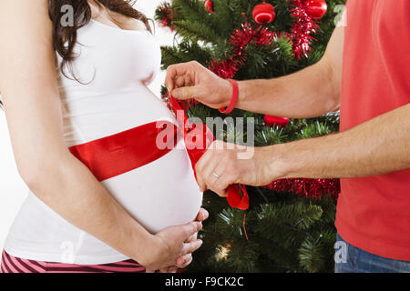 Coppia giovane è in piedi vicino all'albero di natale. La donna è incinta e legato con un nastro rosso. Foto Stock