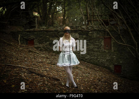 Malice in Wonderland / incubo: una bella ragazza bionda ragazza, con gli occhi bendati, indossando un 'Alice' abito tipo tonaca in piedi da soli di fronte a una casa abbandonati nel deserto tetro bosco foresta, REGNO UNITO Foto Stock