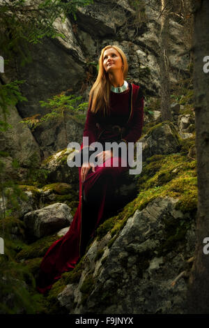 Una donna bellissima fata con lunghi capelli biondi in un abito storico è seduta amids moos rocce coperte in incantevole paesaggio forestral Foto Stock