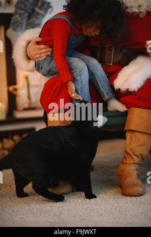 Santa Claus alimentare un gatto con il simpatico bambino africano vicino al camino e albero di Natale a casa Foto Stock