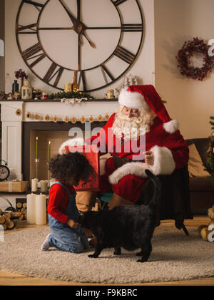 Santa Claus alimentare un gatto con il simpatico bambino africano vicino al camino e albero di Natale a casa Foto Stock