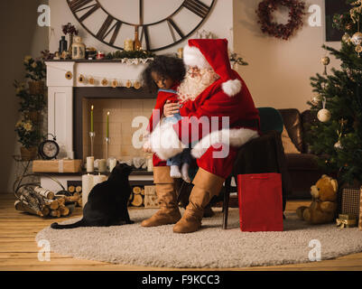 Santa Claus alimentare un gatto con il simpatico bambino africano vicino al camino e albero di Natale a casa Foto Stock