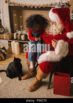 Santa Claus alimentare un gatto con il simpatico bambino africano vicino al camino e albero di Natale a casa Foto Stock