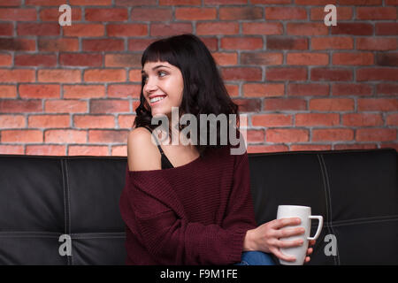 Sorridenti giovane donna in maglia maglione bere il tè sul divano nero su un muro di mattoni sfondo Foto Stock