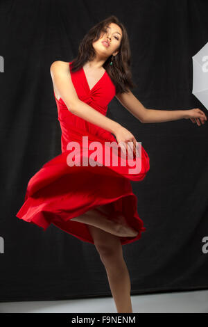 Donna giovane ballerino in abito rosso in una danza moderna girare, con sfondo nero e bianco di piano e di una parte di un ombrello di studio. Foto Stock