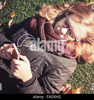 Bionda caucasica ragazza adolescente che stabilisce nel parco autunnali e utilizzo di smart phone con le cuffie in stile vintage tonale filtro di correzione Foto Stock