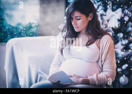 Carino dolce donna incinta seduta vicino al bellissimo albero di Natale decorato a casa e il libro di lettura Foto Stock