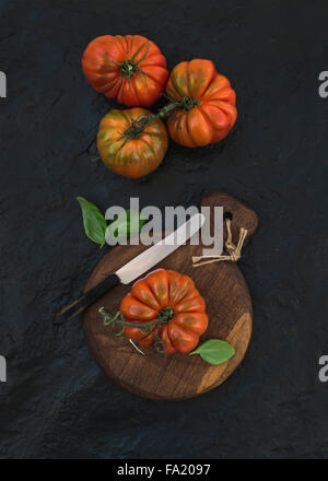 Fresche e mature hairloom i pomodori e le foglie di basilico rustico di legno su pietra nera di sfondo, vista dall'alto Foto Stock