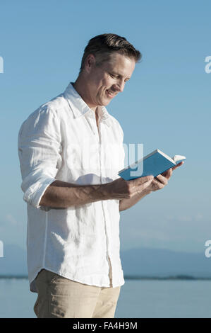 Uomo maturo la lettura del libro e sorridente a Lakeside, Baviera, Germania Foto Stock