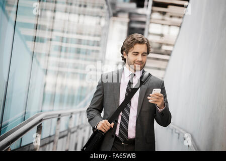 Un uomo che porta una borsa per computer con una fascetta su un passaggio pedonale della città guardando il suo smart phone. Foto Stock