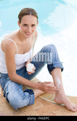 Giovane donna seduta dalla piscina e applicare una lozione solare Foto Stock