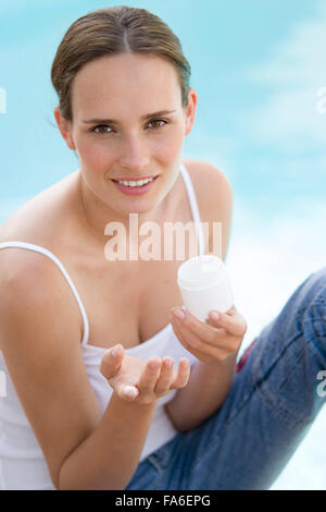 Giovane donna seduta dalla piscina e applicare una lozione solare Foto Stock