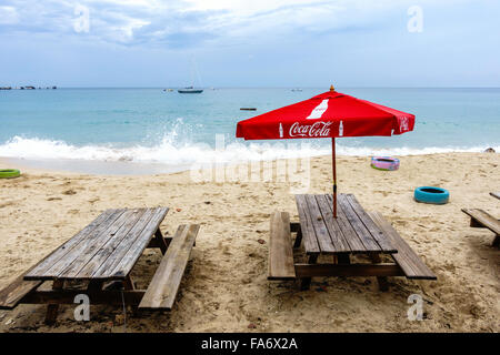 Legno Weatherbeaten tavoli da picnic con un rosso CocaCola ombrellone su una spiaggia caraibica. Foto Stock