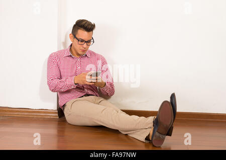 Ispanico maschio rosso che indossa una camicia bianca, khaki jeans seduto sul pavimento di legno contro la parete premendo telefono mobile, sfondo vuoto Foto Stock