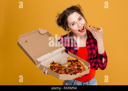 Bella felice giovane donna in maglia a scacchi holding e mangiare la pizza su sfondo giallo Foto Stock