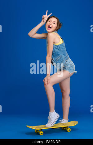 Allegro felice giovane donna in jeans corti e gilet in piedi su skateboard e mostrando segno di vittoria su sfondo blu Foto Stock