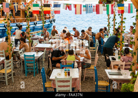 Santorini, Korfos, Cadouni, café, il ristorante sulla spiaggia. Affollate di turisti seduti ai tavoli e sedie per mangiare sotto la fila di mini bandiere nazionali. Foto Stock