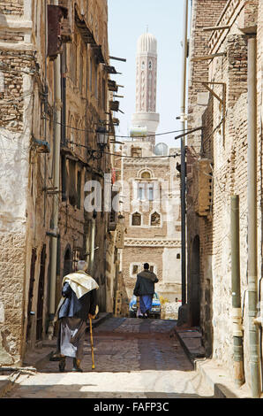 Un uomo con un bastone a camminare per le strade della città vecchia di Sana'a, decorate case, minareto, Yemen, vita quotidiana Foto Stock