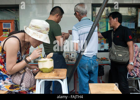 Donna di bere in una noce di cocco nella parte anteriore di un amuleti stallo nella Street a Chinatown di Bangkok, Thailandia. Yaowarat, Bangkok Chinatown, è la più rinomata cucina di strada di destinazione e il locale preferito cenare distretto. Foto Stock
