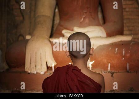 Vista posteriore del giovane monaco novizio pregando a lume di candela all'interno di un tempio buddista, Bagan, Myanmar. Foto Stock
