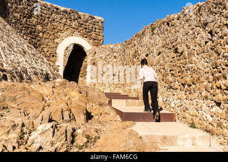 Una donna cammina fino di gradini di pietra accanto a un muro di pietra verso un portale ad arco in un castello. Foto Stock
