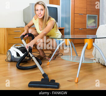 Giovane donna bionda in shorts pulizia con aspirapolvere sul pavimento in parquet a casa Foto Stock