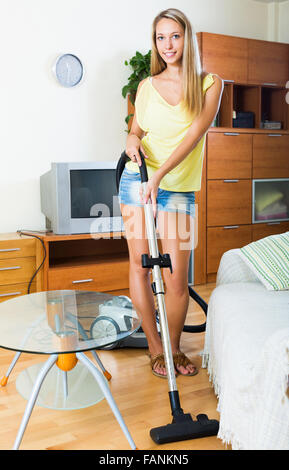 Bionda sorridente ragazza in pantaloncini corti la pulizia con aspirapolvere sul pavimento in parquet a casa Foto Stock