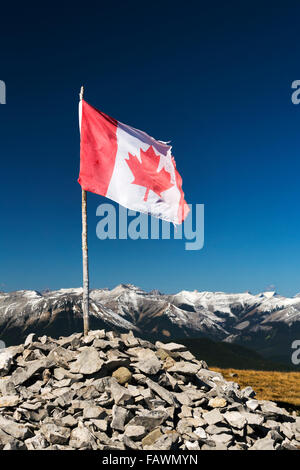 Ben indossati bandiera canadese sul ramo di legno puntellato in una pila di rocce sulla cima di una montagna con la gamma della montagna e cielo blu in background Foto Stock