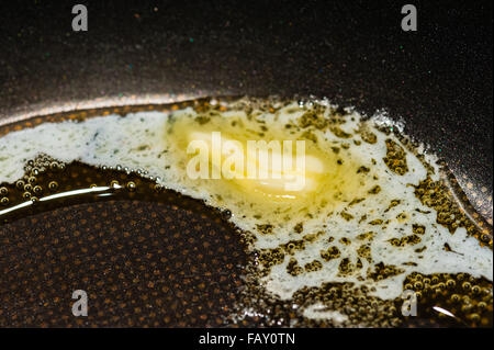 Macro Close-up di un pezzo di burro lentamente la fusione e la formazione di bolle in Teflon nero padella. Foto Stock