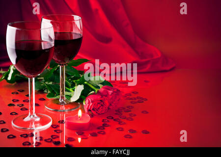 Un romantico ancora in vita con due bicchieri di vino rosso a forma di cuore candela e due rose, con un morbido sfondo satinato. Foto Stock