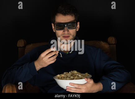 Giovane uomo seduto in camera oscura nella parte anteriore della tv guardare film e mangiare popcorn, indossando occhiali 3D Foto Stock