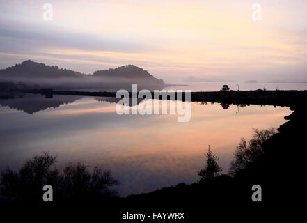 Alba sul lago e colline tagliata da una strada rialzata con bassa giacente nebbia atmosfera di prestito al riflesso rosa e blu Foto Stock