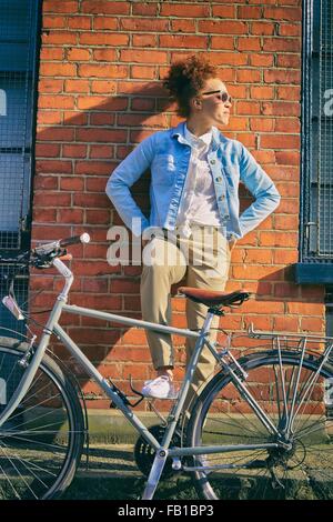 Giovane donna con la bicicletta indossando occhiali da sole appoggiati contro un muro di mattoni, le mani sui fianchi che guarda lontano Foto Stock