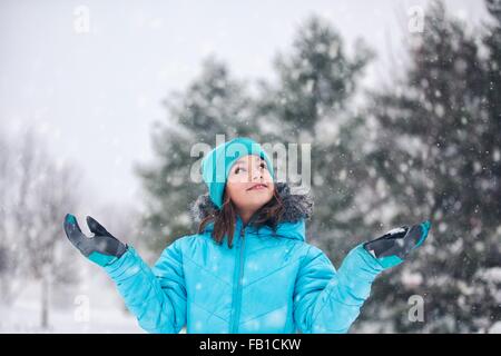 Ragazza indossare un turchese knit hat e ricoprire i bracci sollevati, mani fuori la cattura di neve, cercando di sorridere Foto Stock