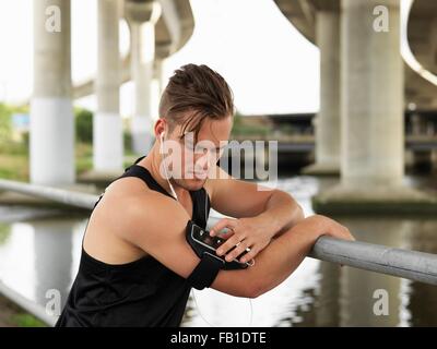 Giovane uomo accanto al fiume, poggiando su ringhiere, utilizzando un lettore MP3 collegato al braccio Foto Stock